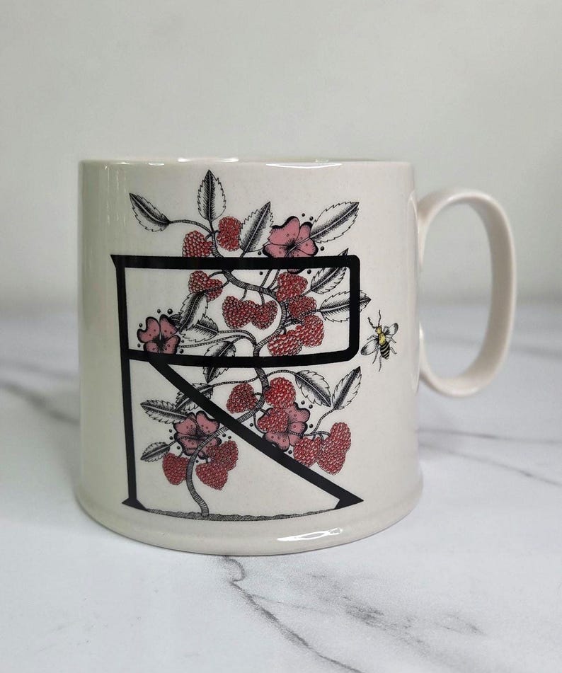 Puede incluir: Taza de cer&aacute;mica color crema con la letra negra &laquo;R&raquo;, adornada con bayas rojas, flores rosas y hojas verdes. Tambi&eacute;n se representa una peque&ntilde;a abeja. La taza tiene un asa curvada.
