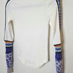 May include: White long-sleeved shirt with a mock turtleneck. The sleeves feature a patterned design with blue, orange, and black accents. The cuffs have a purple leopard print. The shirt has a black stripe down the sides.