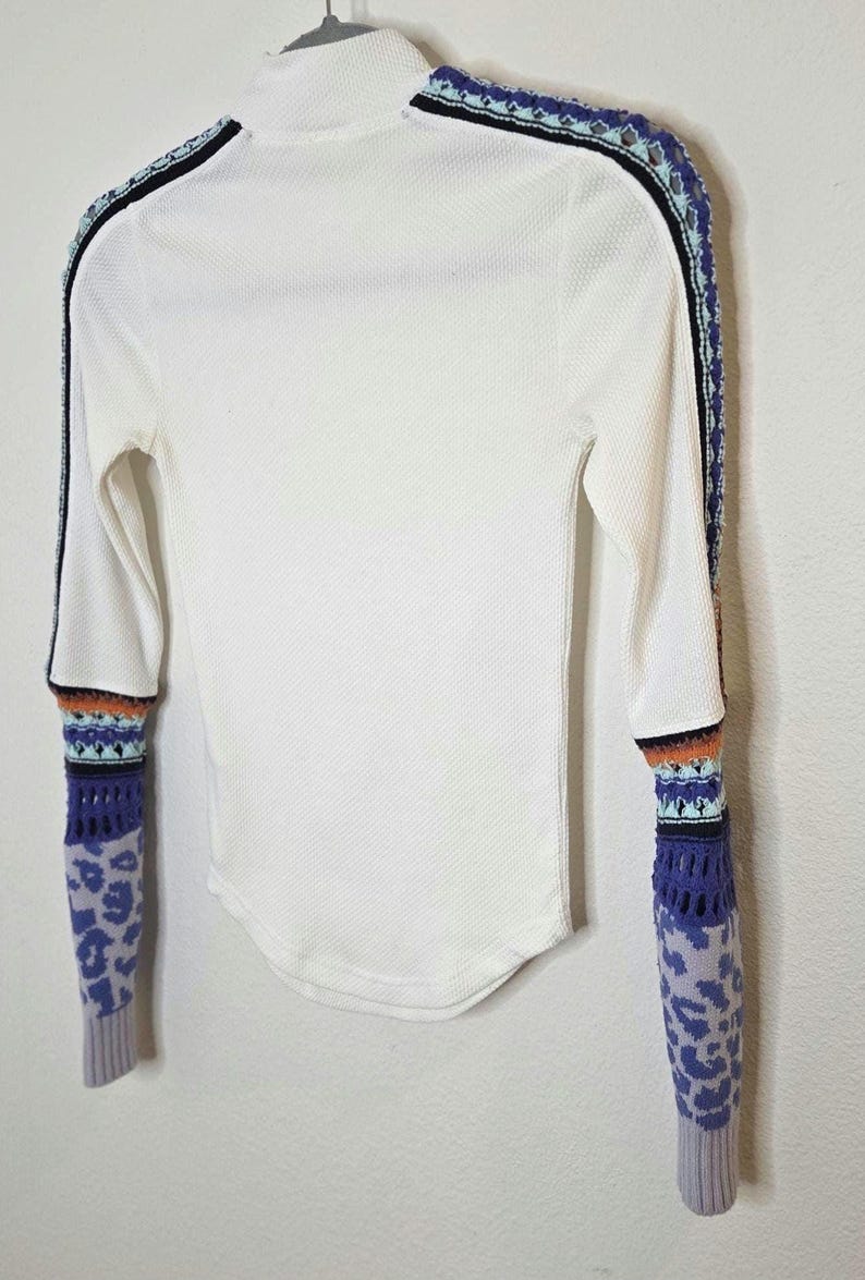May include: White long-sleeved shirt with a mock turtleneck. The sleeves feature a patterned design with blue, orange, and black accents. The cuffs have a purple leopard print. The shirt has a black stripe down the sides.