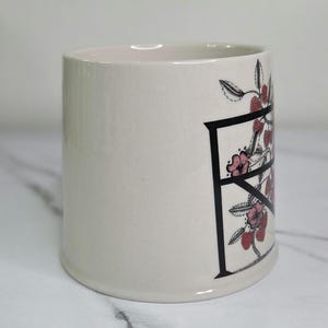 Puede incluir: Una taza de cer&aacute;mica color crema con un dise&ntilde;o de la letra "F" en negro. La letra est&aacute; adornada con flores rosas y bayas rojas. La taza tiene forma cil&iacute;ndrica y un acabado liso y brillante.