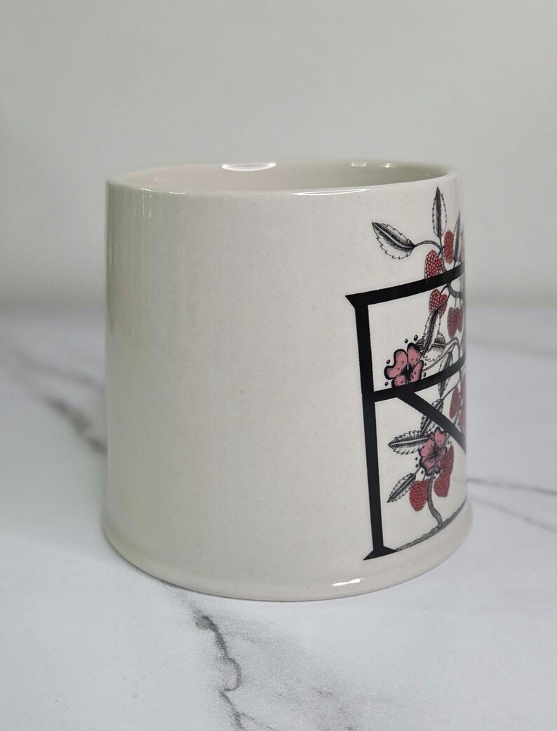 Puede incluir: Una taza de cer&aacute;mica color crema con un dise&ntilde;o de la letra "F" en negro. La letra est&aacute; adornada con flores rosas y bayas rojas. La taza tiene forma cil&iacute;ndrica y un acabado liso y brillante.
