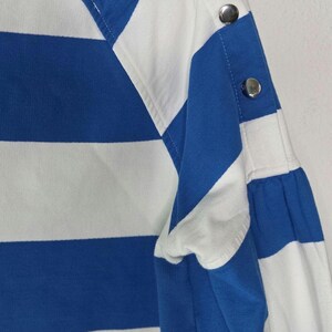 May include: Close-up of a blue and white striped long-sleeved shirt. The shirt features a boat neck and silver button details on the shoulder and sleeve. The fabric has a soft appearance.