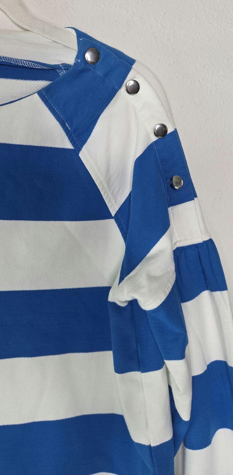 May include: Close-up of a blue and white striped long-sleeved shirt. The shirt features a boat neck and silver button details on the shoulder and sleeve. The fabric has a soft appearance.