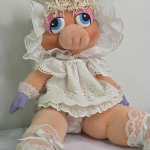May include: A plush pig doll with a pink face, blue eyes, and blonde hair. The doll wears a white lace bonnet, dress, and stockings. The doll's arms are purple, and the doll is sitting down.