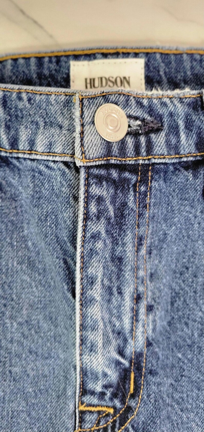 May include: Close-up of blue denim jeans with the brand name "HUDSON" on a white tag. The jeans have a button closure and visible stitching. The denim fabric has a textured appearance.