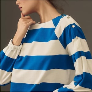 May include: A blue and white striped long-sleeve shirt. The shirt features wide horizontal stripes and silver button details on the shoulder and sleeves. The sleeves have a gathered cuff. The shirt is made of a soft material.