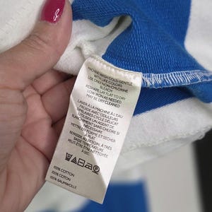 May include: Close-up of a white and blue striped garment. The garment has a blue collar and a tag with care instructions in English and French. The tag indicates the garment is 100% cotton.
