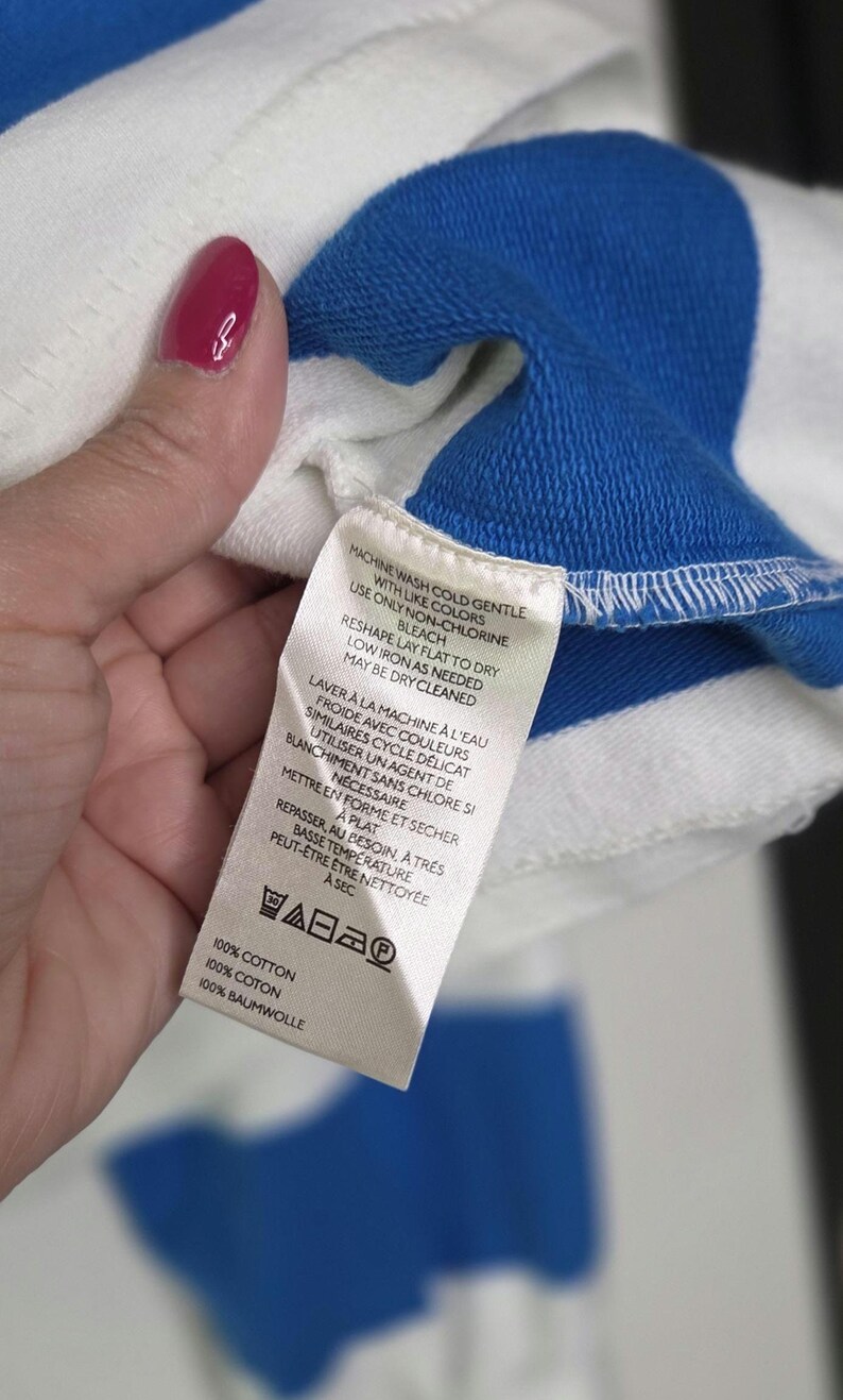 May include: Close-up of a white and blue striped garment. The garment has a blue collar and a tag with care instructions in English and French. The tag indicates the garment is 100% cotton.