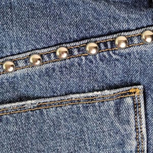 May include: Close-up of a blue denim mini skirt. The skirt features silver-colored studs along the pocket and waistline. The label reads "THE VIPER MINI SKIRT 28".