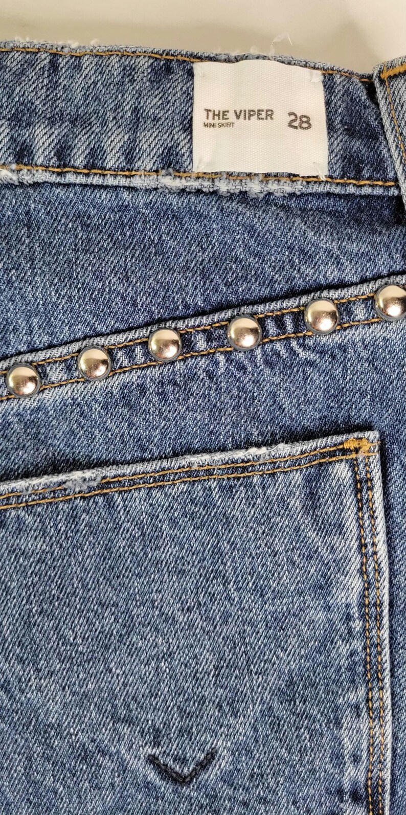 May include: Close-up of a blue denim mini skirt. The skirt features silver-colored studs along the pocket and waistline. The label reads "THE VIPER MINI SKIRT 28".