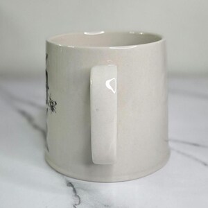 Puede incluir: Una taza de cer&aacute;mica de color crema con un asa blanca. La taza tiene forma cil&iacute;ndrica y un dise&ntilde;o sencillo. El asa es rectangular y est&aacute; sujeta al lateral de la taza. La taza est&aacute; sobre una superficie blanca.