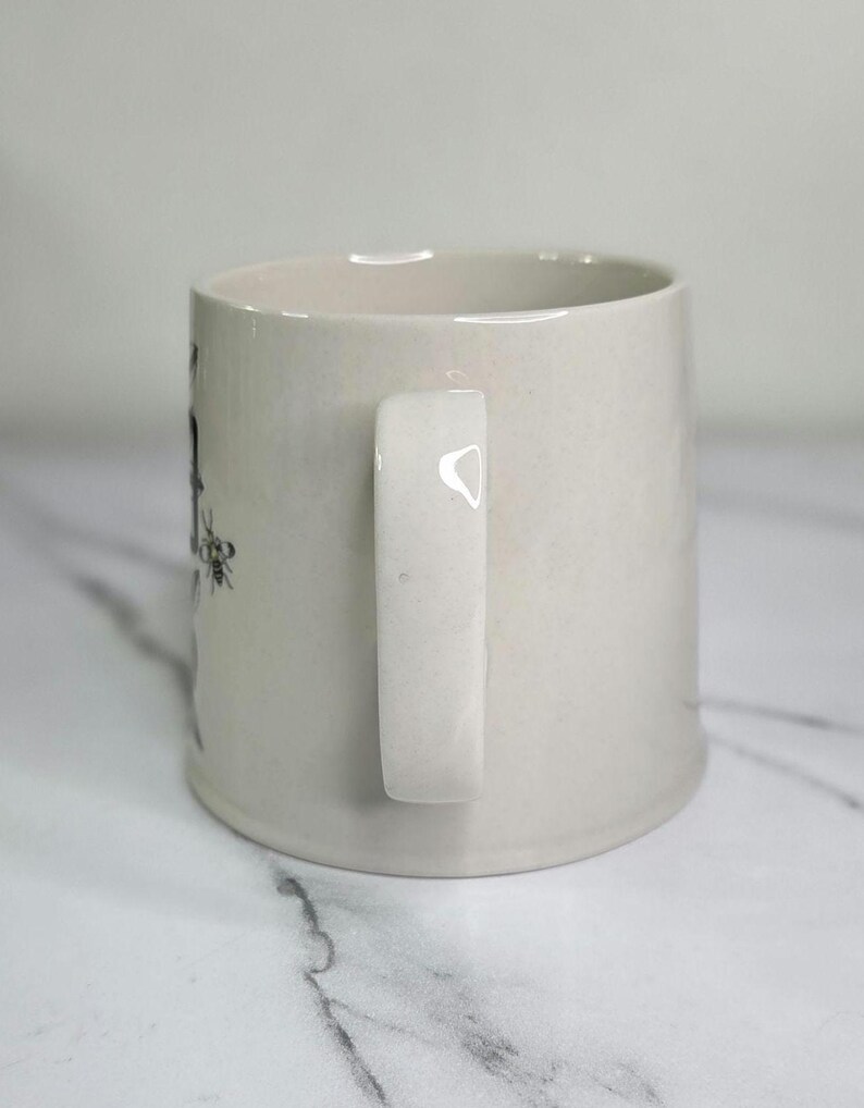 Puede incluir: Una taza de cer&aacute;mica de color crema con un asa blanca. La taza tiene forma cil&iacute;ndrica y un dise&ntilde;o sencillo. El asa es rectangular y est&aacute; sujeta al lateral de la taza. La taza est&aacute; sobre una superficie blanca.