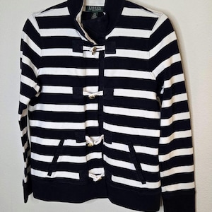 May include: Navy and white striped jacket with toggle closures. The jacket has a high collar, two front pockets, and a black trim on the cuffs and hem. The jacket is hanging on a hanger.