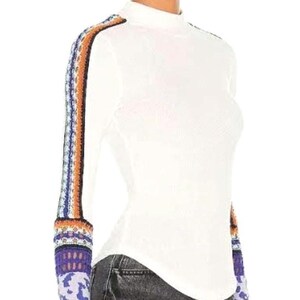 May include: White long-sleeve top with a mock turtleneck. The sleeves feature a colorful, patterned design in shades of blue, orange, and purple. The top is paired with dark gray jeans.