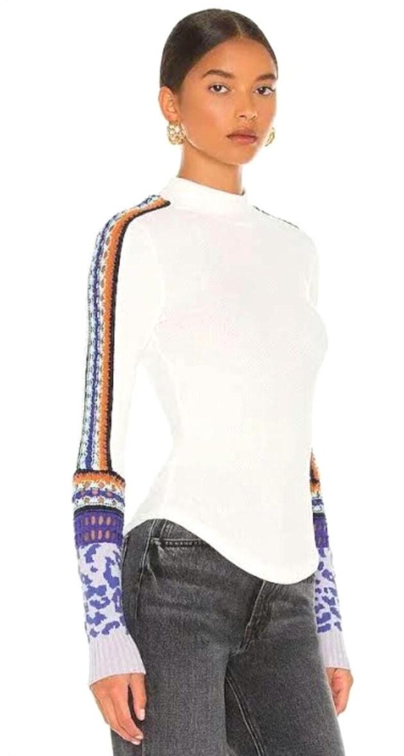 May include: White long-sleeve top with a mock turtleneck. The sleeves feature a colorful, patterned design in shades of blue, orange, and purple. The top is paired with dark gray jeans.