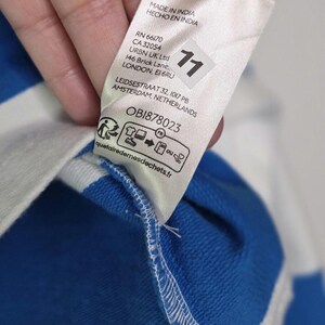 May include: Close-up of a white tag with text in English and Spanish, including "100% COTONE" and "MADE IN INDIA." The tag is attached to a blue and white striped fabric. A number "11" is printed on the tag.
