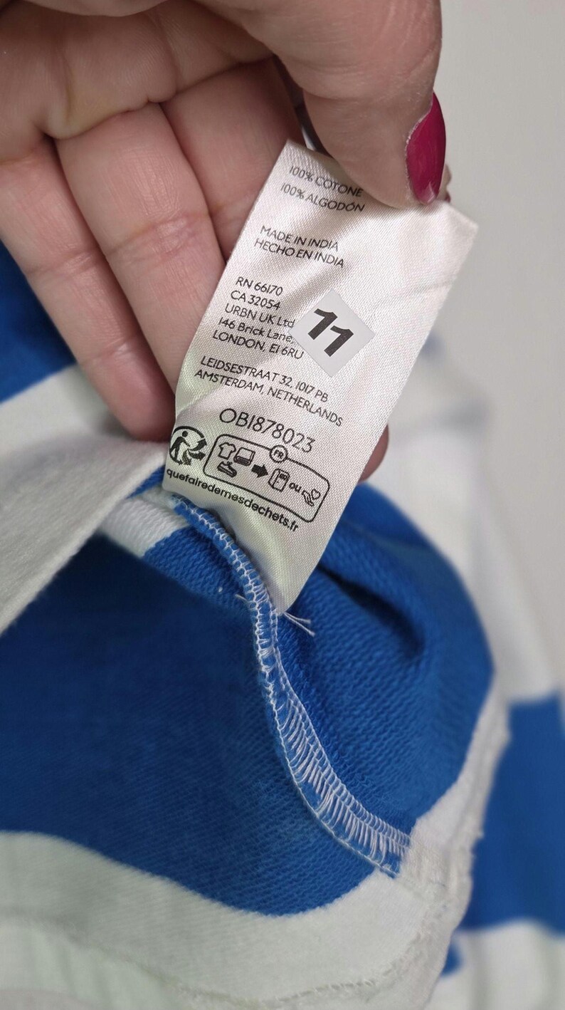 May include: Close-up of a white tag with text in English and Spanish, including "100% COTONE" and "MADE IN INDIA." The tag is attached to a blue and white striped fabric. A number "11" is printed on the tag.