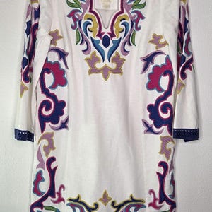 May include: White tunic top with colorful embroidered floral designs in pink, purple, blue, and yellow. The tunic has three-quarter sleeves and a V-neckline. The hem and sleeve cuffs are trimmed with a navy blue border.