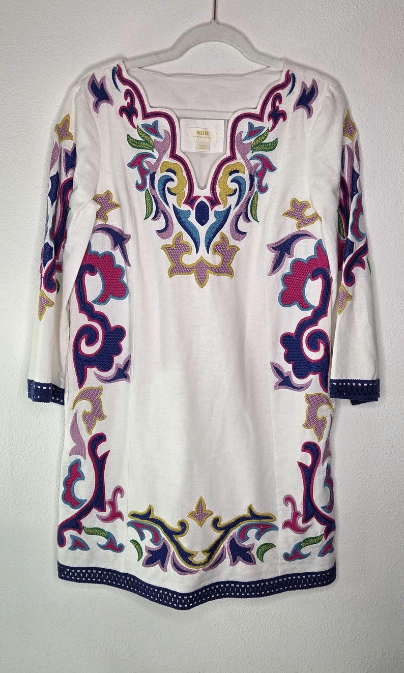 May include: White tunic top with colorful embroidered floral designs in pink, purple, blue, and yellow. The tunic has three-quarter sleeves and a V-neckline. The hem and sleeve cuffs are trimmed with a navy blue border.