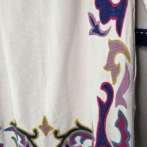 May include: White garment with intricate embroidered floral patterns in shades of blue, pink, purple, and green. The edges are trimmed with a dark blue crocheted border. The design is a close-up of the garment's details.