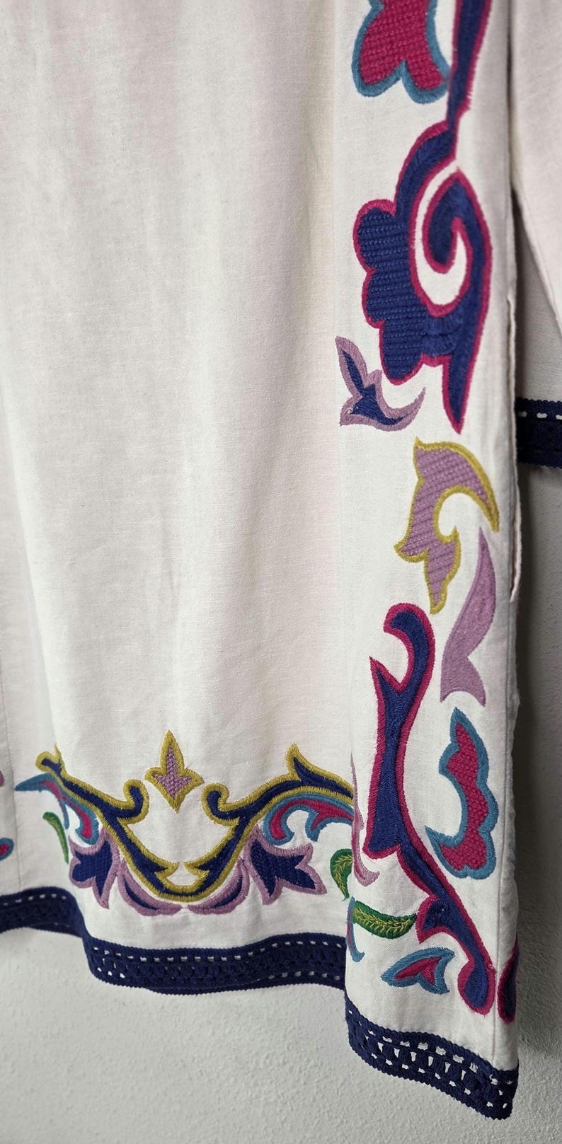 May include: White garment with intricate embroidered floral patterns in shades of blue, pink, purple, and green. The edges are trimmed with a dark blue crocheted border. The design is a close-up of the garment's details.