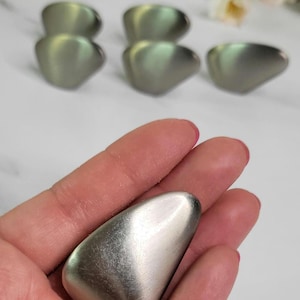 May include: A collection of silver-toned, teardrop-shaped objects. One is held in a hand, while others are arranged in the background. The objects have a smooth, metallic finish, reflecting light. The overall aesthetic is minimalist and modern.