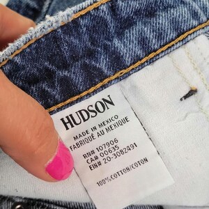 May include: Close-up of blue denim jeans with a white tag. The tag reads "HUDSON" and includes text such as "MADE IN MEXICO" and "100% COTTON". The jeans have metal rivets and a button closure.