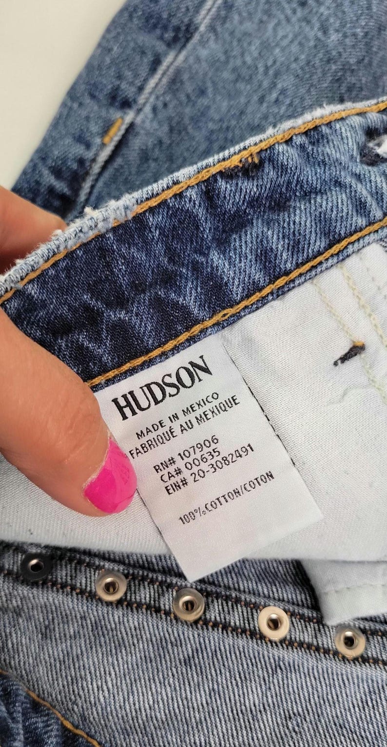 May include: Close-up of blue denim jeans with a white tag. The tag reads "HUDSON" and includes text such as "MADE IN MEXICO" and "100% COTTON". The jeans have metal rivets and a button closure.