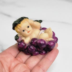 May include: A small figurine of a baby lying in a cluster of purple grapes. The baby has blonde hair, a white diaper, and is surrounded by green leaves. The figurine is held in a hand.