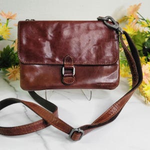 May include: A brown leather crossbody bag with a flap closure and adjustable strap. The bag features a buckle detail on the front and a metal clasp for the strap. The bag is displayed against a backdrop of flowers.