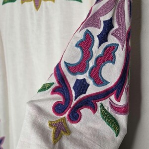 May include: White long-sleeved garment with intricate embroidery in shades of blue, pink, purple, and yellow. The embroidery features floral and geometric patterns, with a focus on the sleeves and neckline.