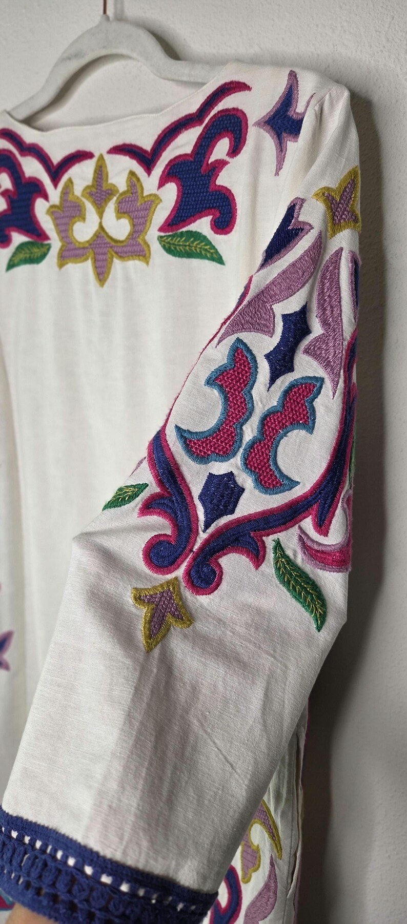 May include: White long-sleeved garment with intricate embroidery in shades of blue, pink, purple, and yellow. The embroidery features floral and geometric patterns, with a focus on the sleeves and neckline.