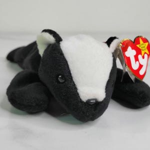 May include: A plush toy badger with a black and white design. The badger has a white face and a black body, with a red heart-shaped tag attached to its ear. The toy is lying down.
