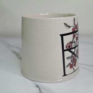 Puede incluir: Taza de cer&aacute;mica color crema con la letra negra "F" decorada con flores rosas y bayas rojas. La taza cil&iacute;ndrica tiene una superficie lisa. El dise&ntilde;o est&aacute; en el lateral de la taza.