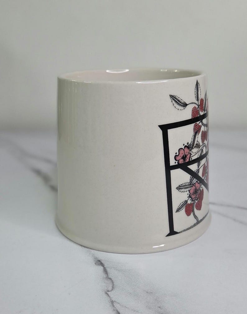 Puede incluir: Taza de cer&aacute;mica color crema con la letra negra "F" decorada con flores rosas y bayas rojas. La taza cil&iacute;ndrica tiene una superficie lisa. El dise&ntilde;o est&aacute; en el lateral de la taza.