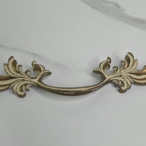 May include: Ornate, antique-style drawer pull with a curved handle. The metal hardware features a distressed, off-white finish on the decorative floral elements and a bronze-colored handle. This decorative hardware is suitable for cabinets and drawers.
