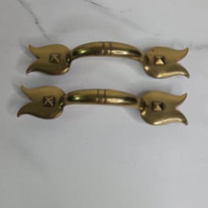 May include: Two brass-toned cabinet pulls with a tulip-shaped design on each end. The pulls have a rectangular center section with two horizontal lines. The ends feature a square design.
