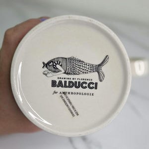Puede incluir: Plato de cer&aacute;mica blanco con una ilustraci&oacute;n de pez en blanco y negro. El plato presenta el texto "BALDUCCI for ANTHROPOLOGIE" y "DRAWING BY FLORENCE". Apto para lavavajillas y microondas.