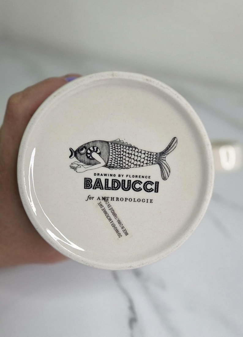 Puede incluir: Plato de cer&aacute;mica blanco con una ilustraci&oacute;n de pez en blanco y negro. El plato presenta el texto "BALDUCCI for ANTHROPOLOGIE" y "DRAWING BY FLORENCE". Apto para lavavajillas y microondas.