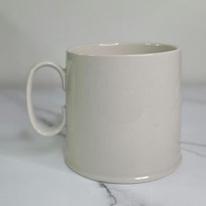 Puede incluir: Una taza de cer&aacute;mica beige claro con un asa redondeada. La taza tiene forma cil&iacute;ndrica y un acabado liso y brillante. La taza est&aacute; sobre una superficie de m&aacute;rmol blanco.