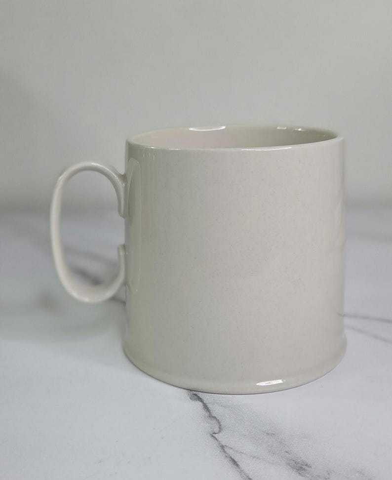 Puede incluir: Una taza de cer&aacute;mica beige claro con un asa redondeada. La taza tiene forma cil&iacute;ndrica y un acabado liso y brillante. La taza est&aacute; sobre una superficie de m&aacute;rmol blanco.