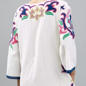 May include: White tunic dress with colorful floral embroidery. The dress features a V-neck and three-quarter sleeves. The embroidery includes pink, blue, green, and yellow accents. The sleeves and hem are trimmed with blue fabric.