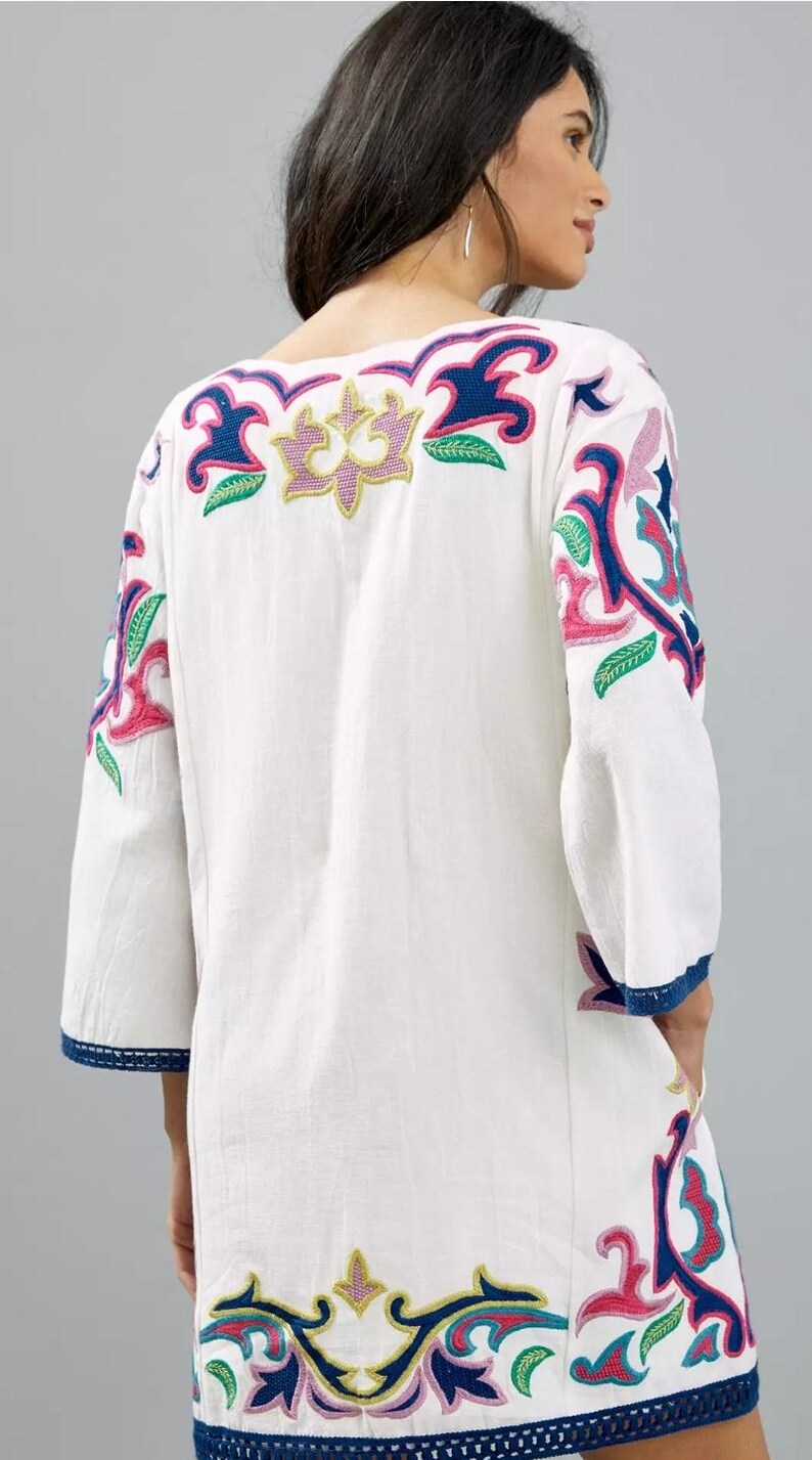May include: White tunic dress with colorful floral embroidery. The dress features a V-neck and three-quarter sleeves. The embroidery includes pink, blue, green, and yellow accents. The sleeves and hem are trimmed with blue fabric.