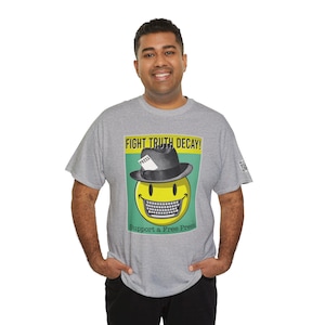 Puede incluir: Una camiseta gris con un gráfico de una cara sonriente que lleva un fedora con la palabra "PRESS" en él. El gráfico también incluye el texto "FIGHT TRUTH DECAY!" y "Support a Free Press".