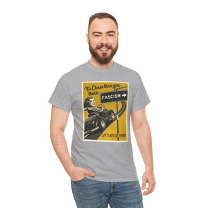 Puede incluir: Una camiseta gris con un gráfico de estilo vintage de una mujer conduciendo un coche hacia un cartel que dice "FASCISMO". El texto en el gráfico dice "¡Está más cerca de lo que piensas! No vayamos allí."