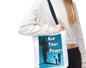 Puede incluir: Bolso tote blanco con una correa azul marino. El bolso presenta un gráfico con la silueta de una persona sosteniendo una herramienta, con las palabras "Use Your Power" en blanco. Debajo, siluetas de dos personas sosteniendo carteles.