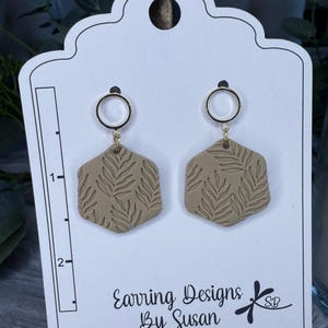 May include: A pair of beige polymer clay earrings with a leaf pattern. The earrings are attached to gold hoops.