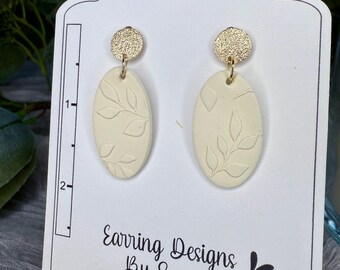 May include: A pair of cream-colored oval earrings with a leaf design. The earrings have gold-colored tops.
