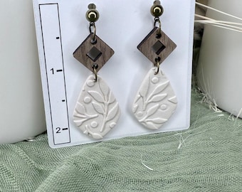 May include: A pair of handmade earrings with a wooden diamond-shaped top and a white teardrop-shaped bottom. The white portion features an embossed leaf design. The earrings are displayed on a white card with the text "Earring Designs By Sue".