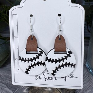 May include: A pair of white and black baseball-themed earrings with brown leather accents. The earrings are on a white card with the text "By Susan" and a signature.